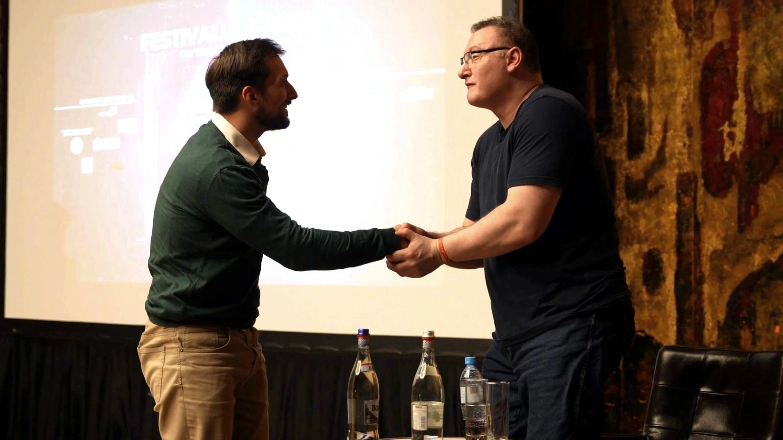 Two people shake hands in front of a screen